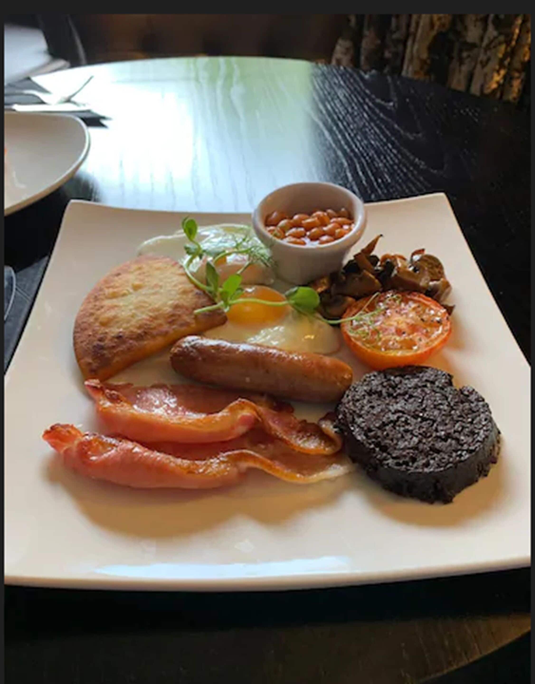 En morgenmadstallerken med æg, bacon, pølser, en skive tomat og en lille skål bønner.