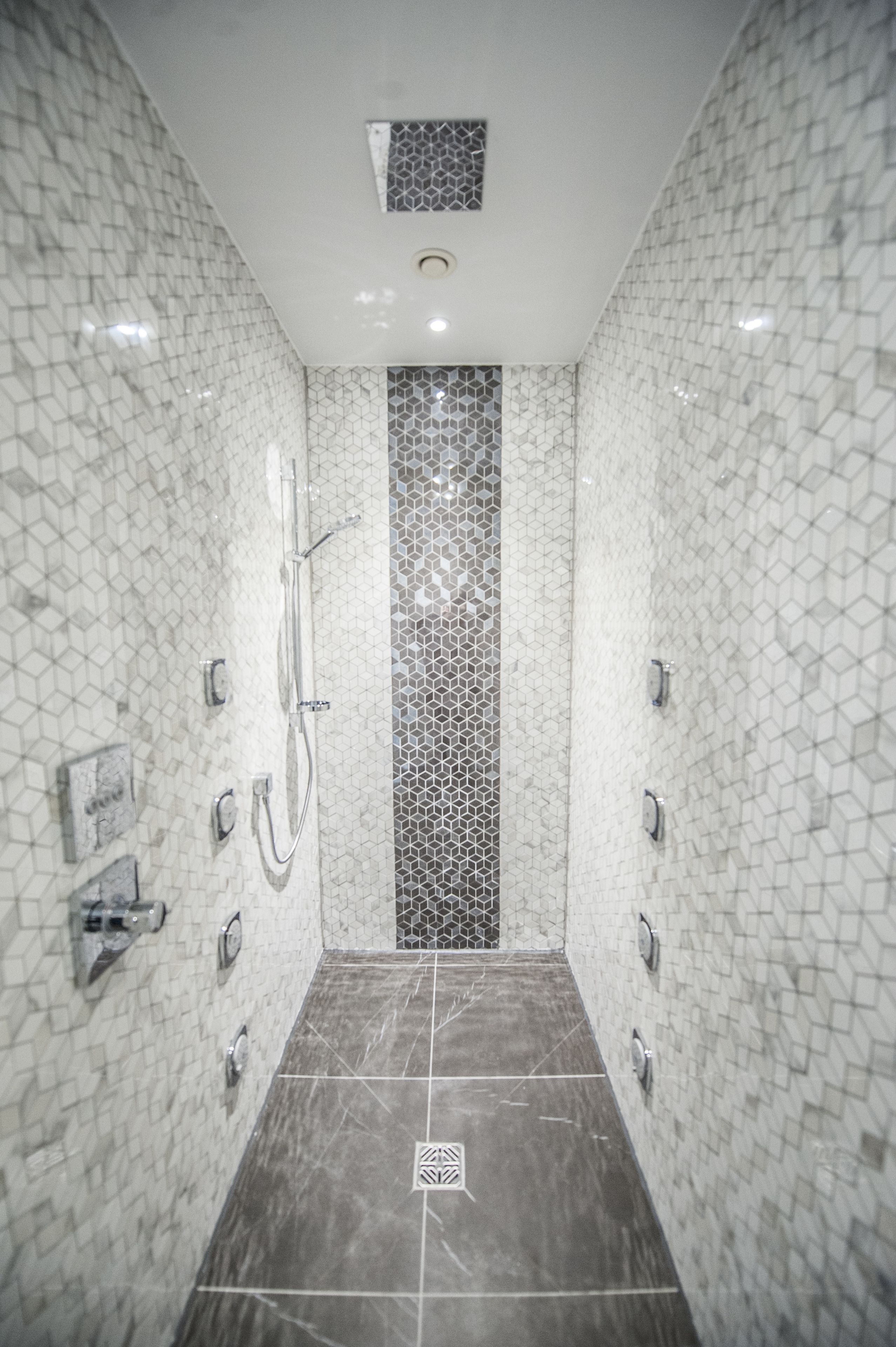 Une douche moderne avec un carrelage aux motifs géométriques, un panneau de commande central et un sol foncé.