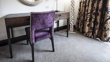 Une chambre d’hôtel avec un bureau en bois, une chaise violette, un miroir rond, une lampe et des rideaux à motifs.