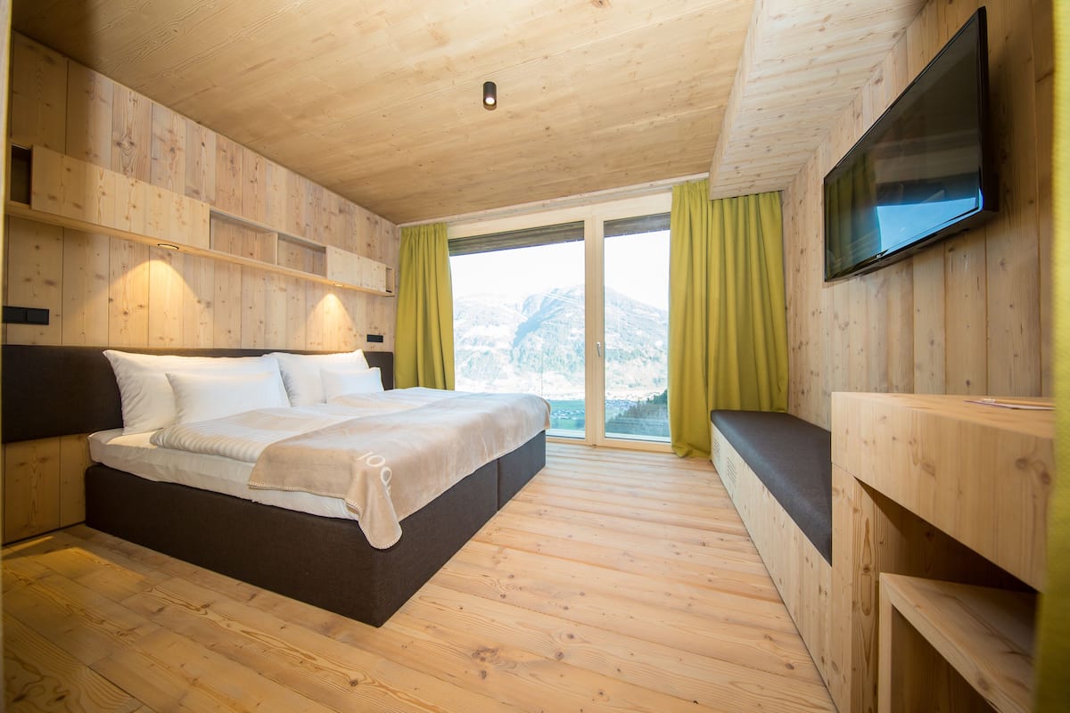 A wooden-paneled bedroom with a bed, a TV mounted on the wall, and a view of mountains through a large window.