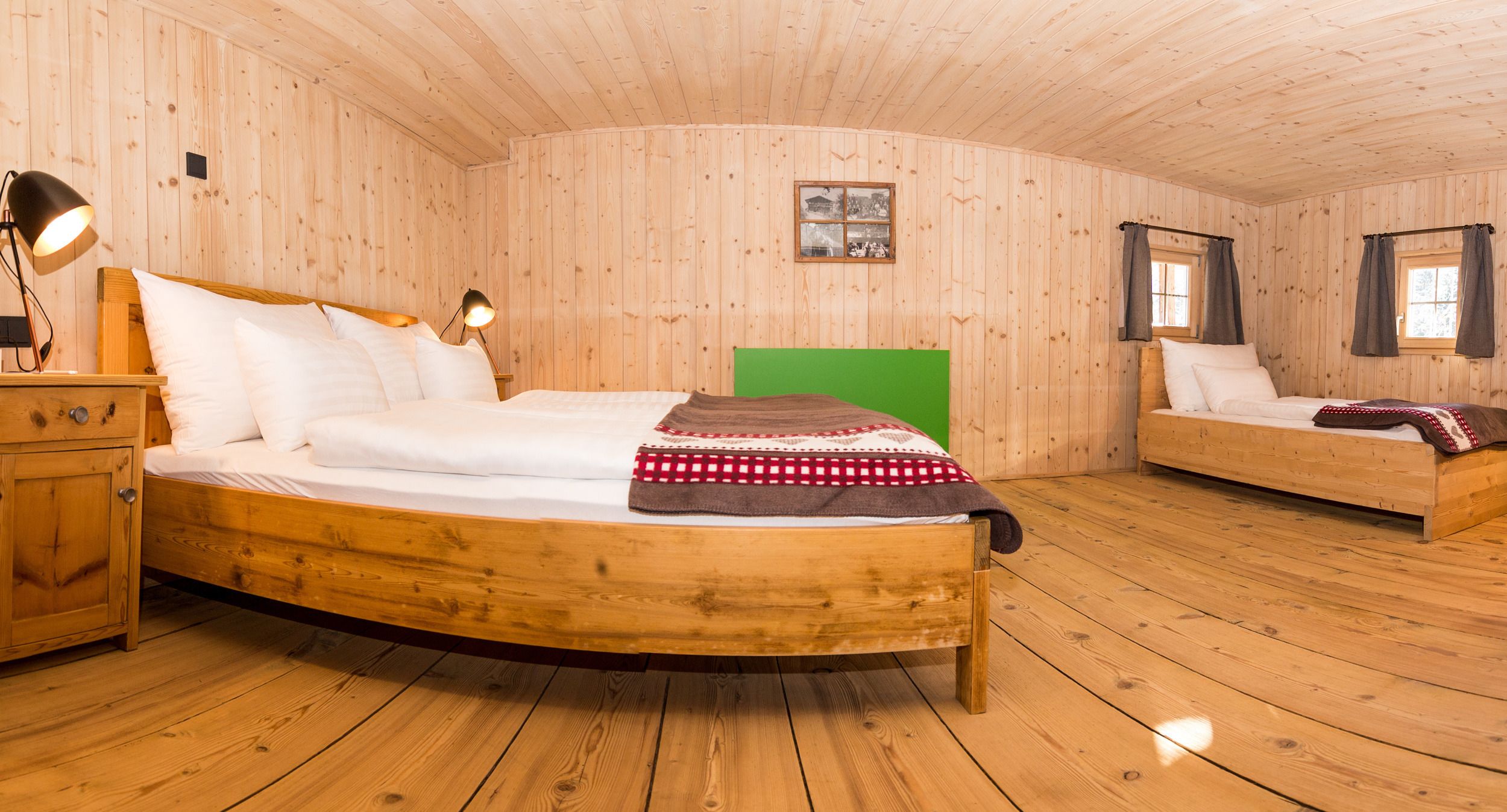 A wooden cabin bedroom with a large bed, a smaller bed, a nightstand, a lamp, and a framed picture on the wall.