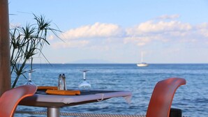 A table set with a wine glass, salt and pepper shakers, and a menu on a table by the sea with a sailboat in the background. - Camping Merendella (San-Nicolao)