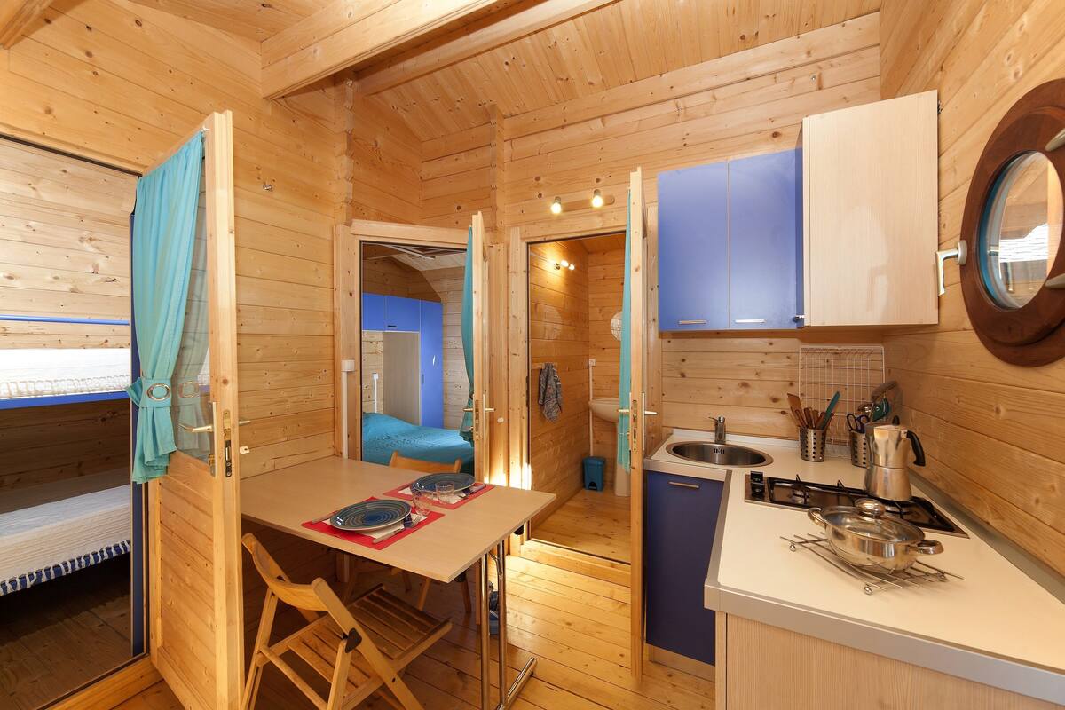 Une petite cabane en bois comprenant un coin repas, une petite cuisine avec un poêle et un espace nuit équipé d’un lit superposé.