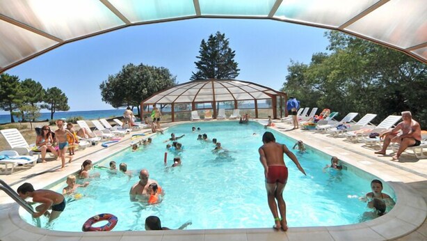 A swimming pool with people swimming and lounging, surrounded by sunbeds and a glass-enclosed structure. - Camping Merendella (San-Nicolao)