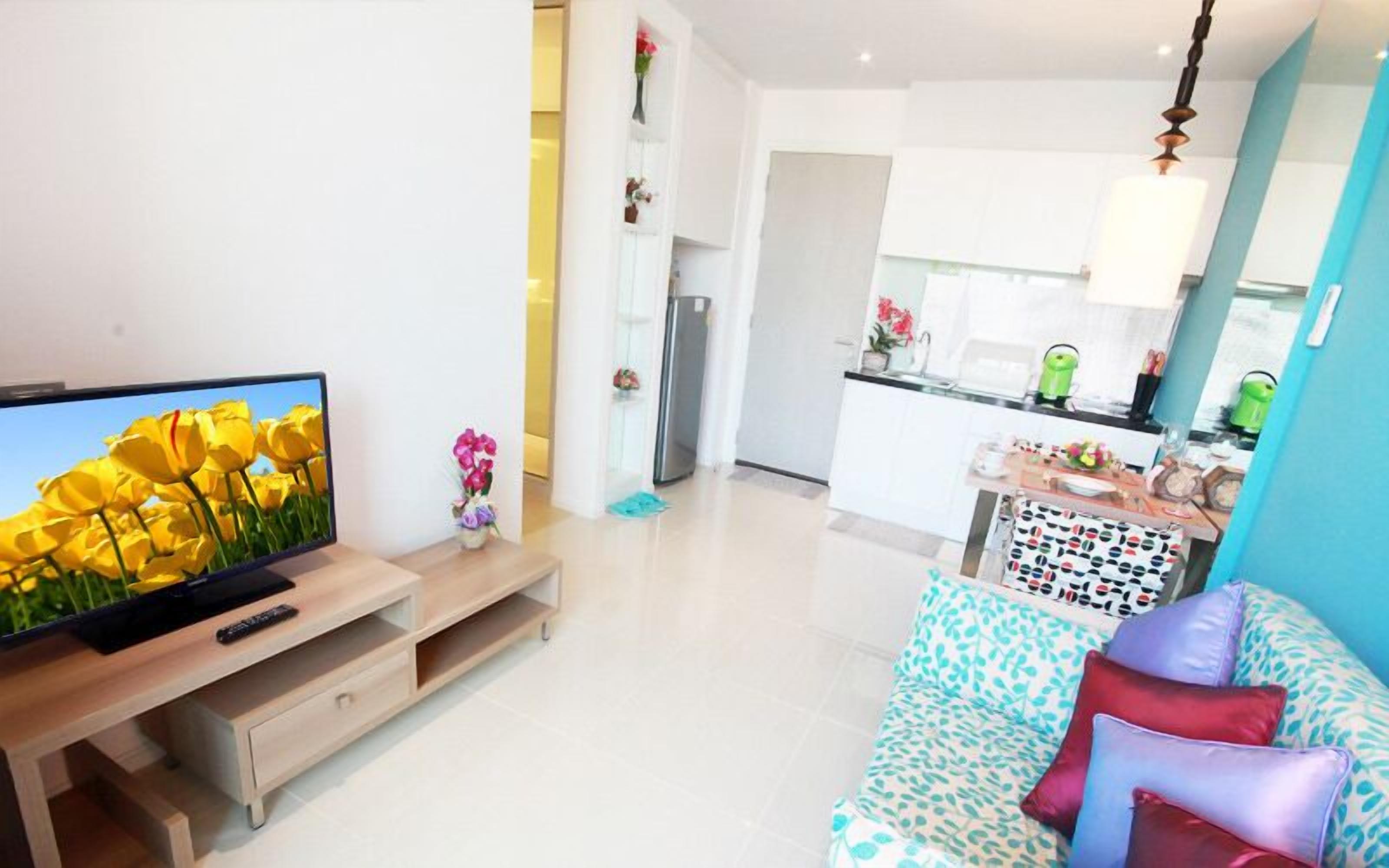 Ein modernes Wohnzimmer mit Fernseher, einem Holztisch, einer Couch mit gemusterten Kissen und einer Küche im Hintergrund.