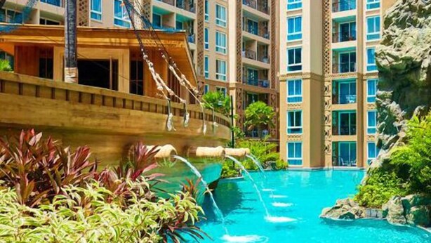 Une piscine dont la structure est en forme de navire, entourée d’une végétation luxuriante et avec un immeuble à plusieurs étages en arrière-plan.