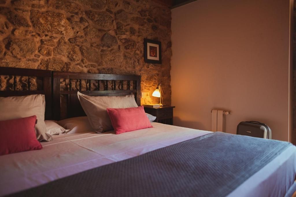 Una cama con pared de piedra de fondo, una mesita de noche con lámpara y una maleta.