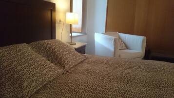 Una habitación de hotel con una cama grande, una mesita de noche con lámpara y una silla con cojín.