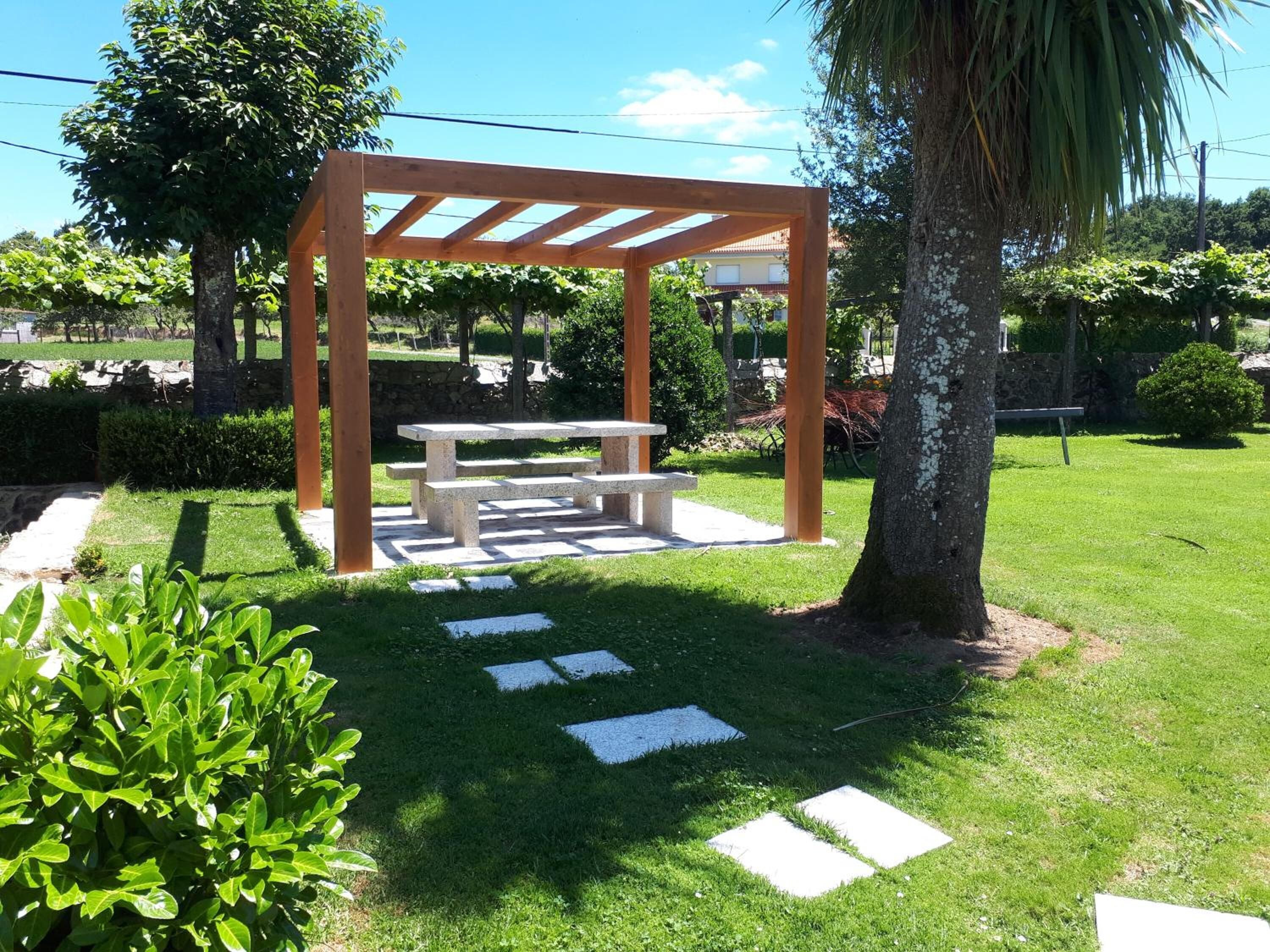Una pérgola de madera con un banco, rodeada de un césped y jardín bien cuidados.