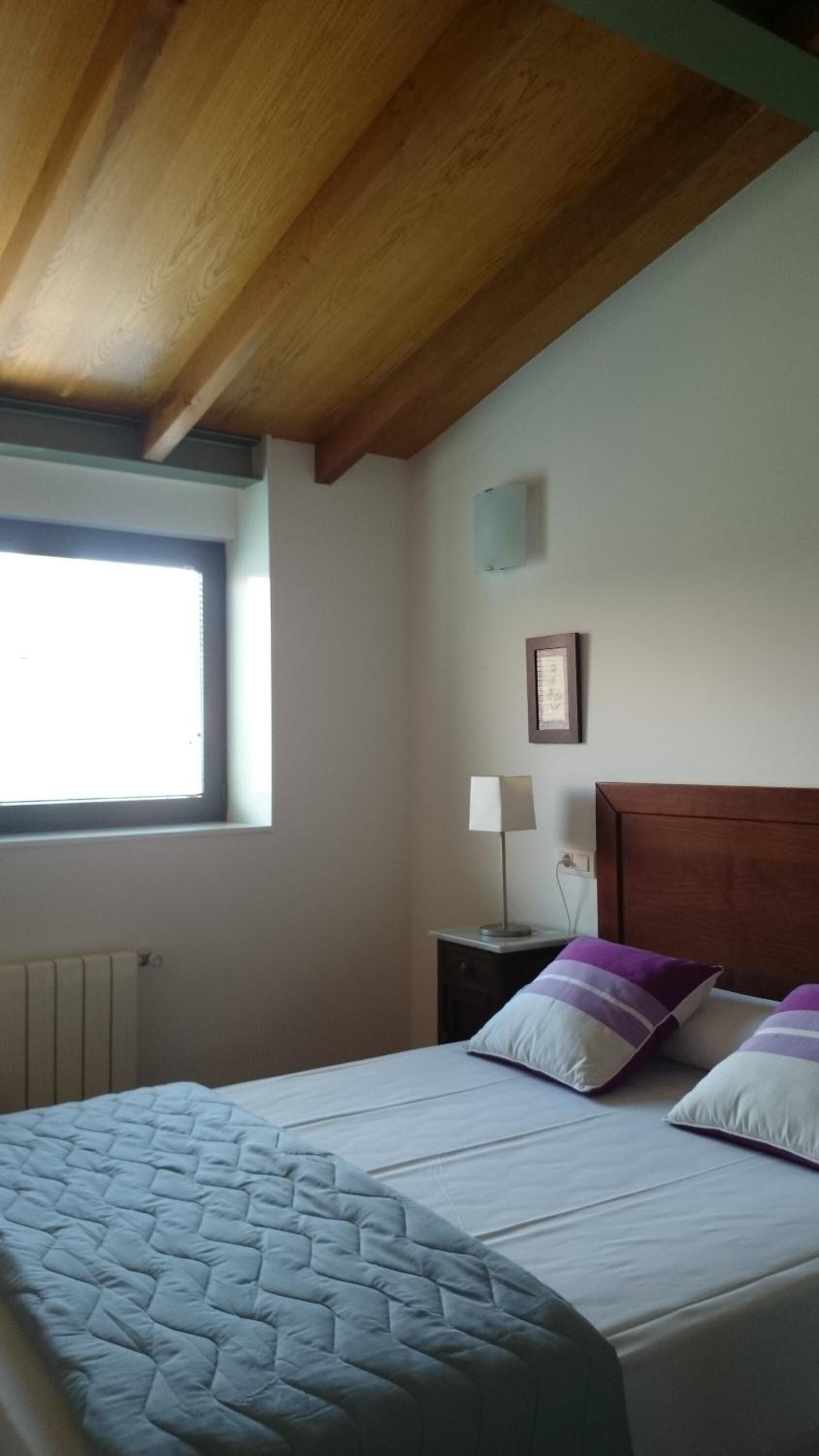 A bedroom with a bed, a window, a nightstand, a lamp, and a framed picture on the wall.