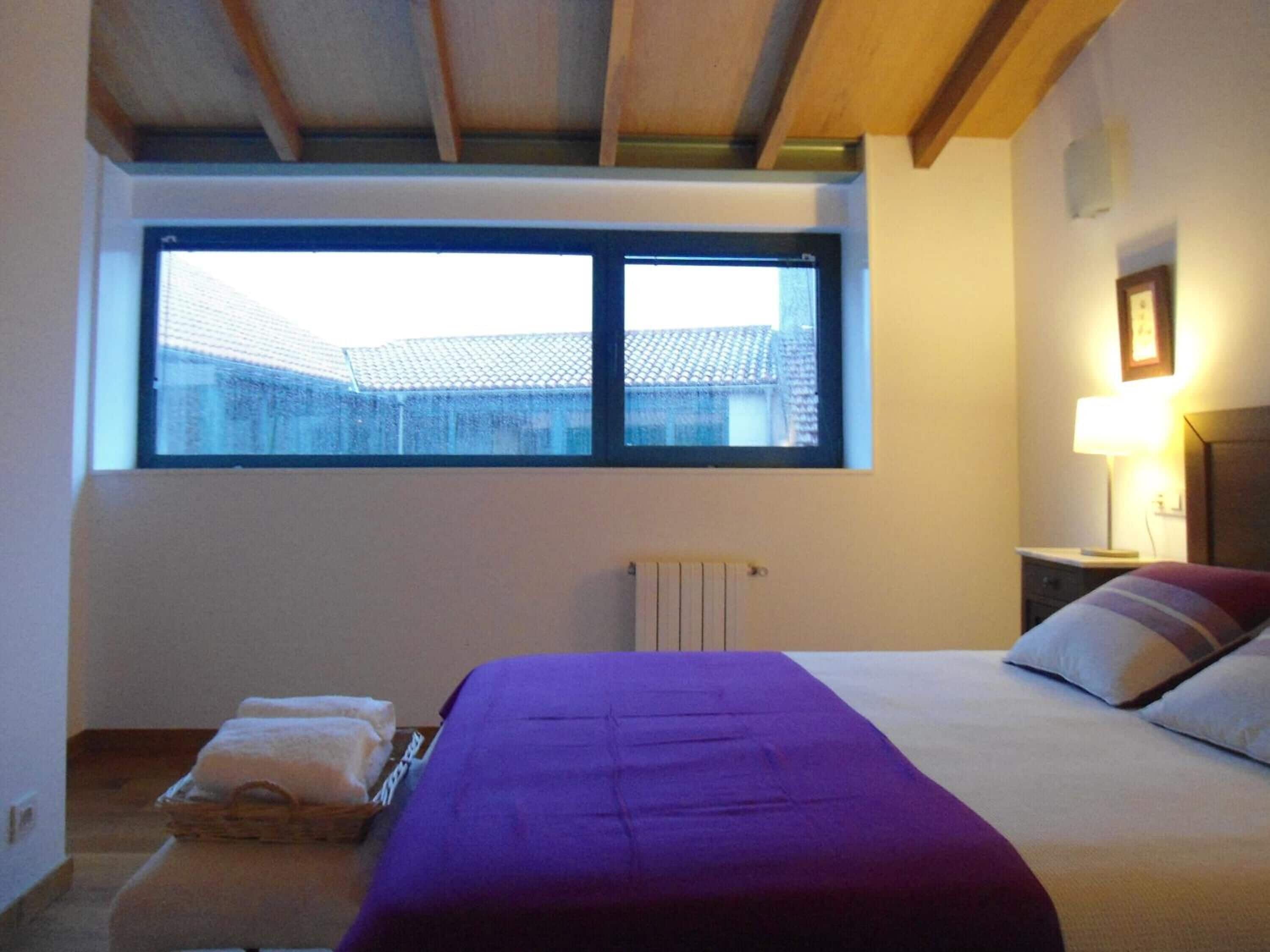 A bedroom with a large window, a bed with a purple bedspread, a bedside lamp, and a radiator.