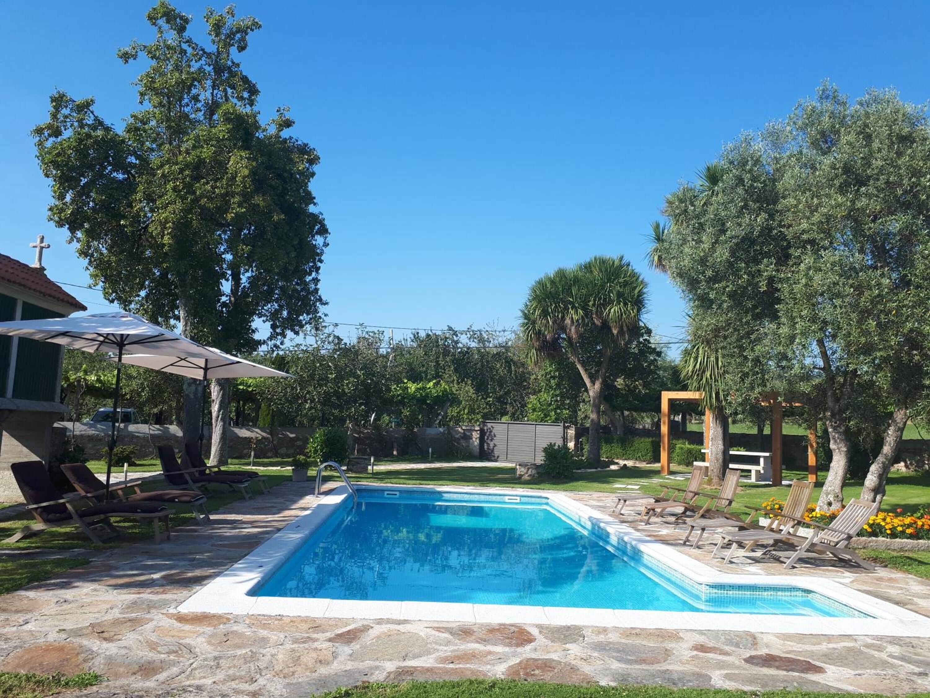 A rectangular swimming pool with a stone border, surrounded by lounge chairs and a covered umbrella.