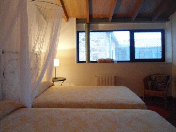 A bedroom with two beds, a window with a view of buildings, a bedside table with a lamp, and a chair.