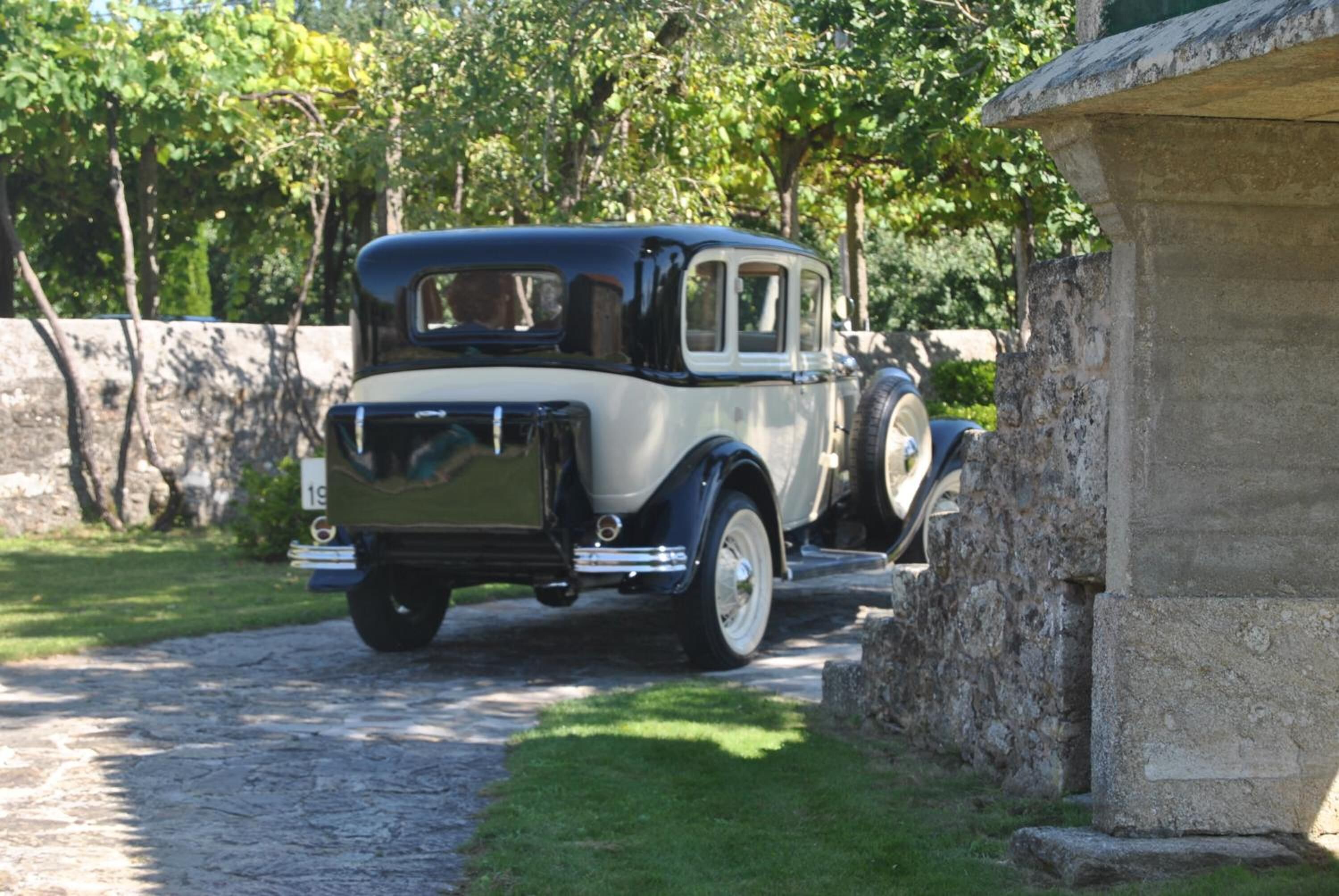 A vintage car parked on a cobblestone path beside a stone wall.