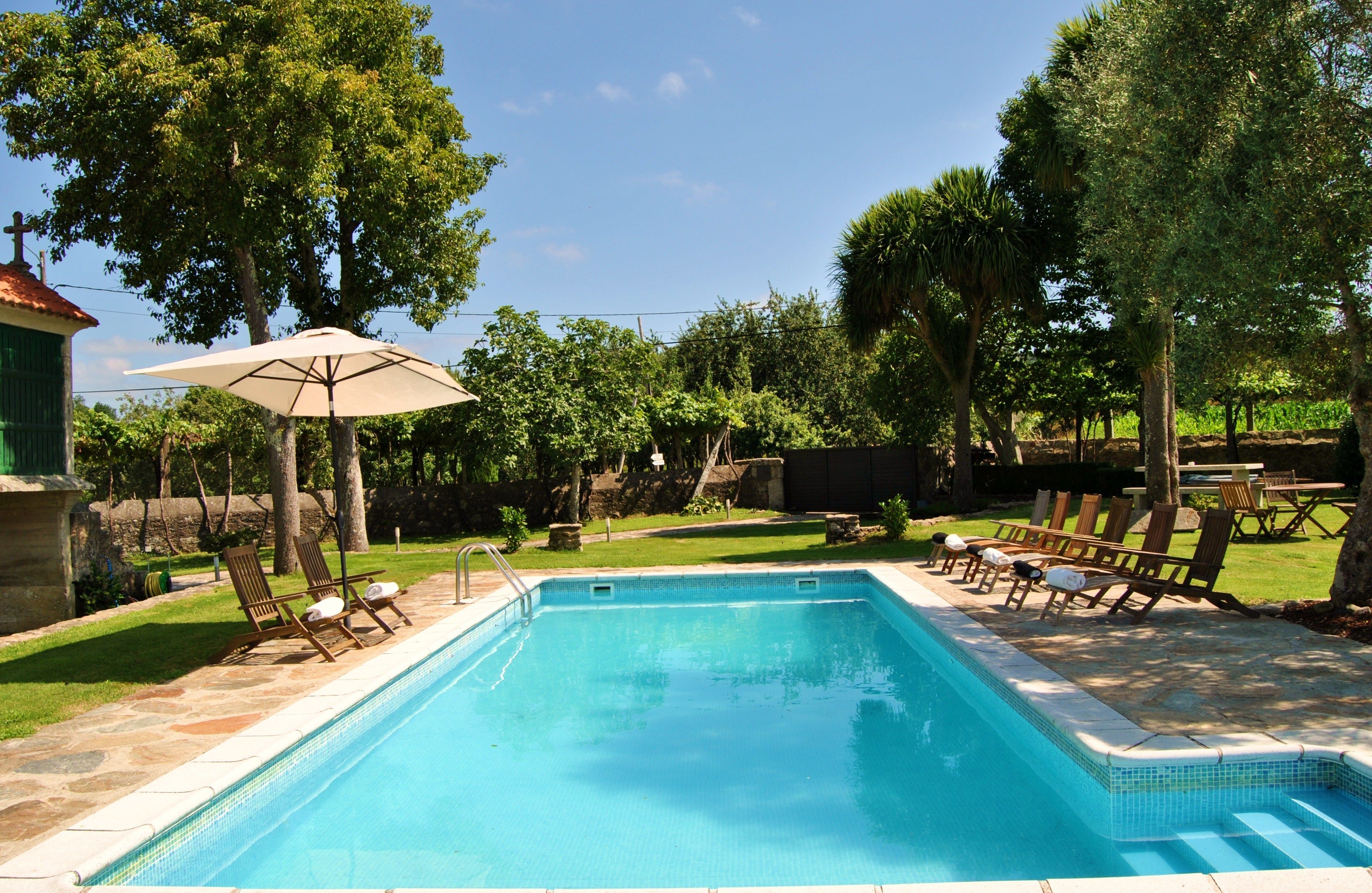 Una piscina rectangular con borde de azulejos, rodeada por una terraza de piedra. Hay sillas reclinables de madera y un paraguas en la terraza.