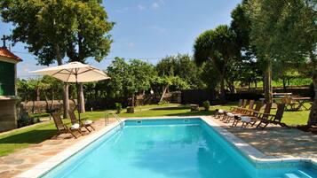 Una piscina rectangular con borde de azulejos, rodeada por una terraza de piedra. Hay sillas reclinables de madera y un paraguas en la terraza.