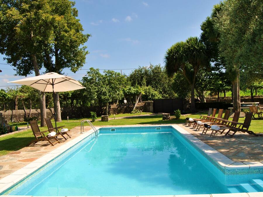 Una piscina rectangular con borde de azulejos, rodeada por una terraza de piedra. Hay sillas reclinables de madera y un paraguas en la terraza.
