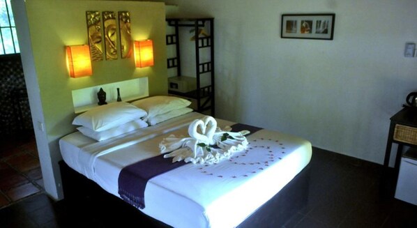 A neatly made bed with white linens, a purple throw blanket, and a decorative swan made of towels. There are wall-mounted lights and a framed picture above the bed.