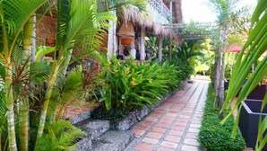 A pathway lined with lush tropical plants and a thatched-roof structure, likely a part of a resort or hotel. - Raingsey Bungalow (Kep)