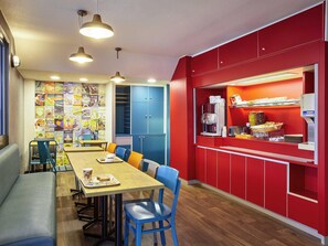 Un petit coin repas avec une table en bois, des chaises bleues et un banc. On y trouve des meubles bleus et un comptoir rouge avec une machine à café et des en-cas.