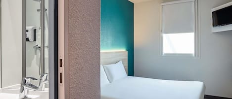 A hotel room with a bed, a white wall, a teal accent wall, a window, a TV mounted on the wall, and a bathroom with a mirror, sink, and shower.