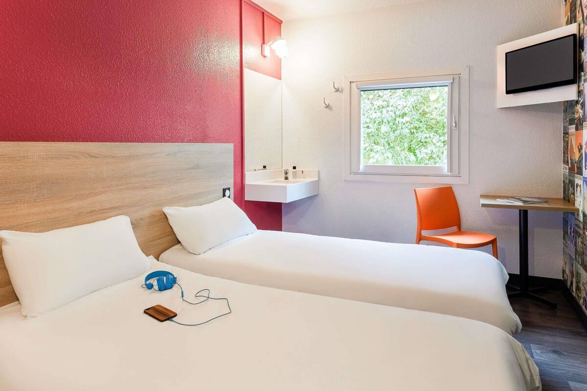 Une chambre d’hôtel avec deux lits, un bureau, une chaise, une télévision et une fenêtre donnant sur un espace verdoyant.