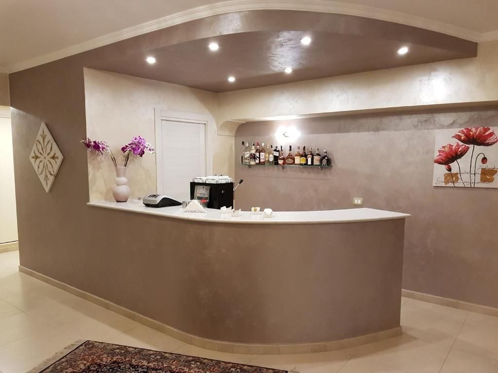 A reception area with a curved counter, a coffee machine, a vase with flowers, and a wall-mounted shelf with bottles.