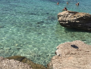 Un plan d’eau peu profond et limpide, avec des rochers visibles et quelques personnes qui se baignent.