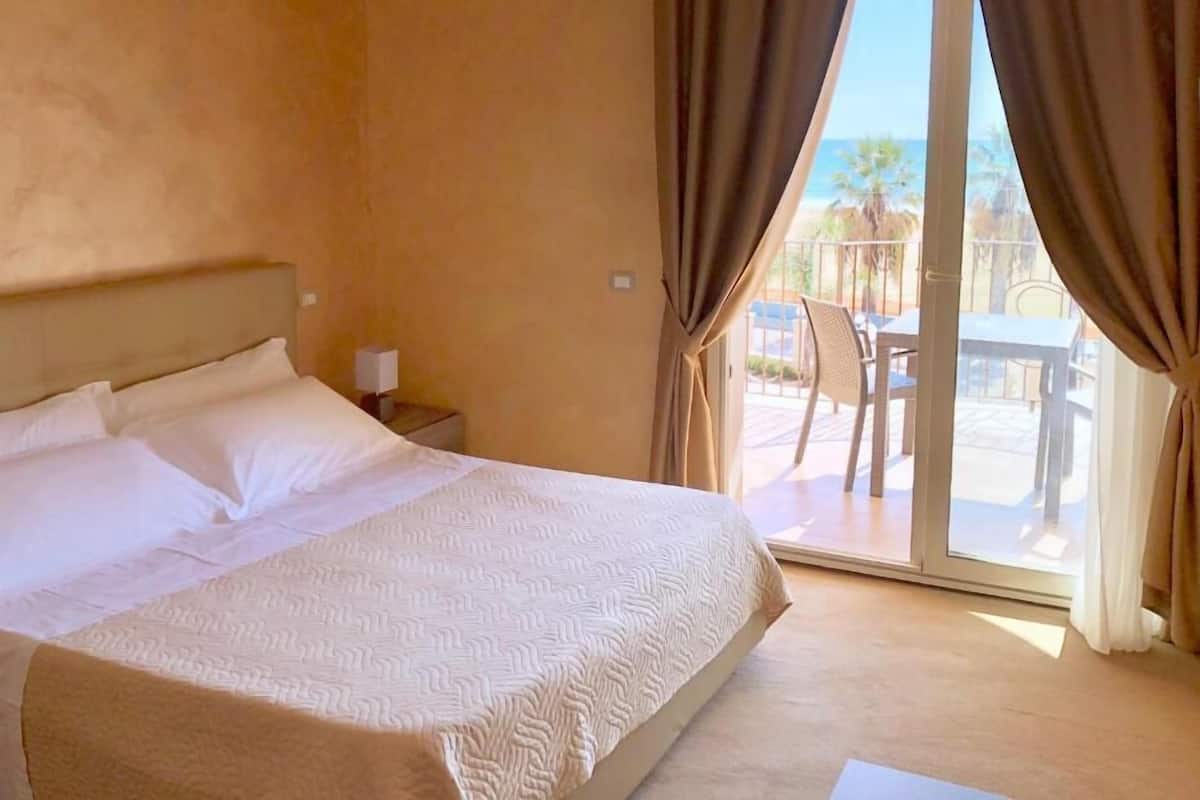 A hotel room with a large bed, a nightstand, and a view of an outdoor patio with a table and chairs.