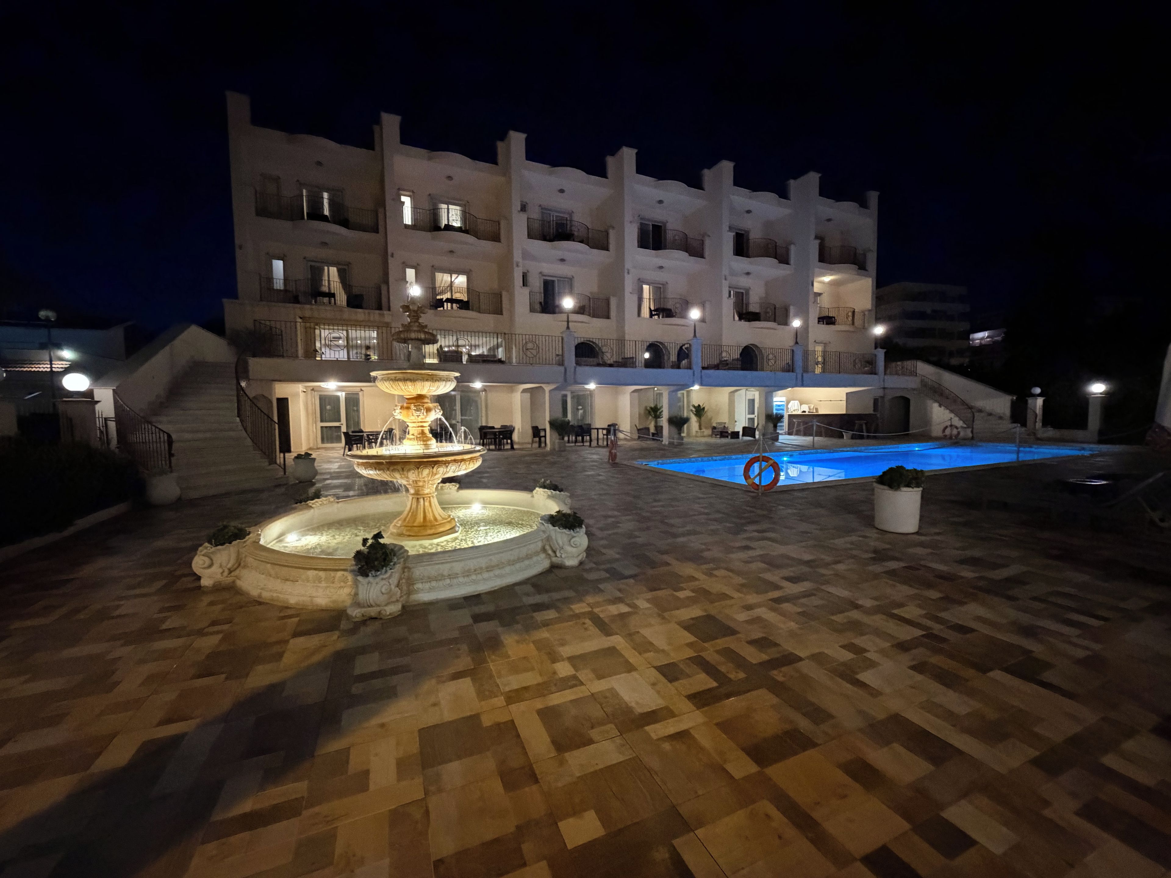 Une scène nocturne représentant la cour d’un hôtel, avec une fontaine centrale, une piscine et un immeuble à plusieurs étages doté de balcons.