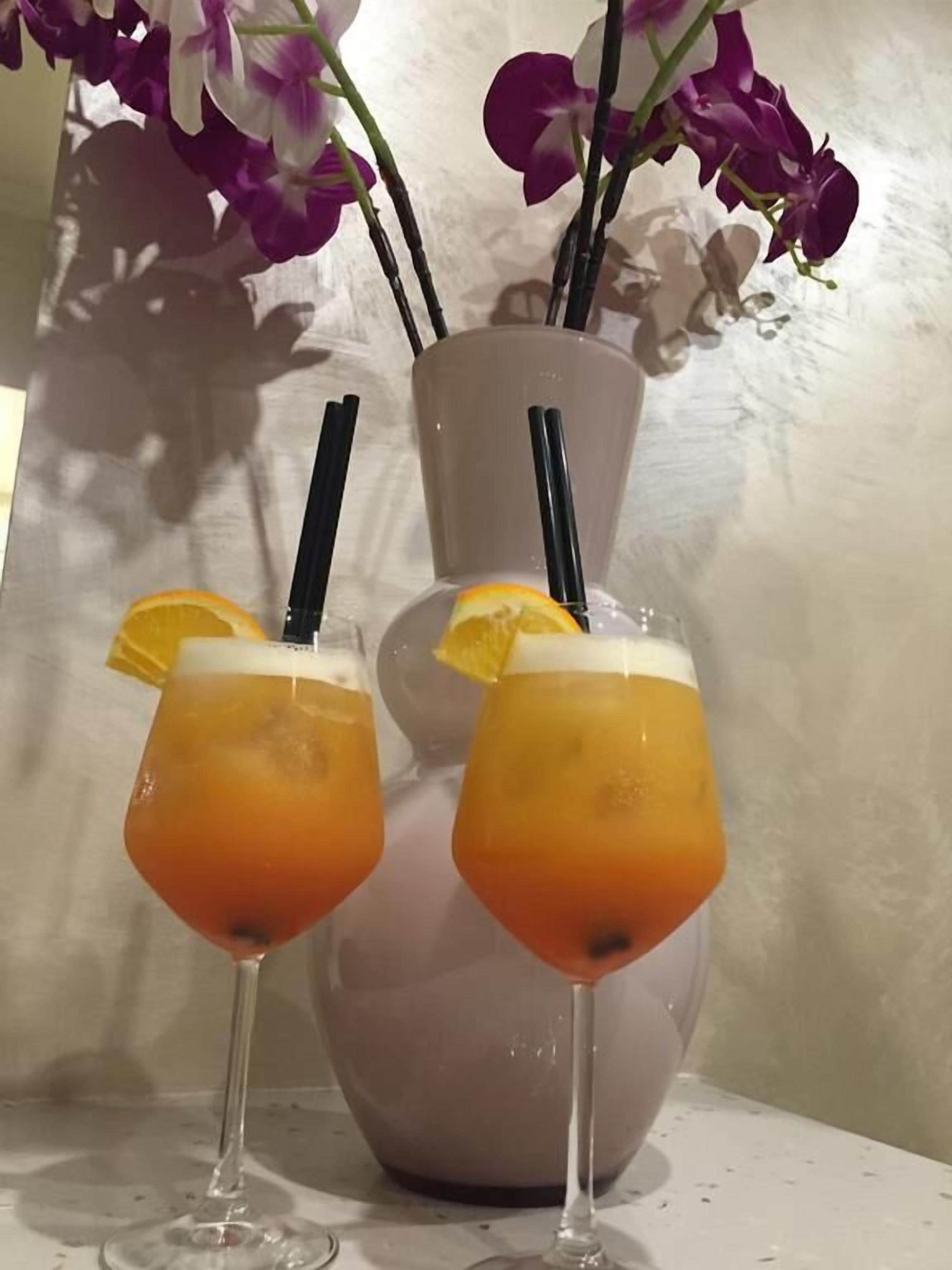 Two glasses of orange-colored beverage with straws and orange slices, placed on a table with a vase of purple flowers in the background.