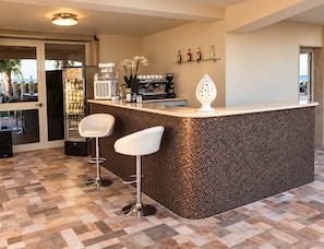 A modern kitchen with a bar area, two bar stools, a coffee machine, and a wine cooler. - Hotel Riviera Palace (Porto Empedocle)