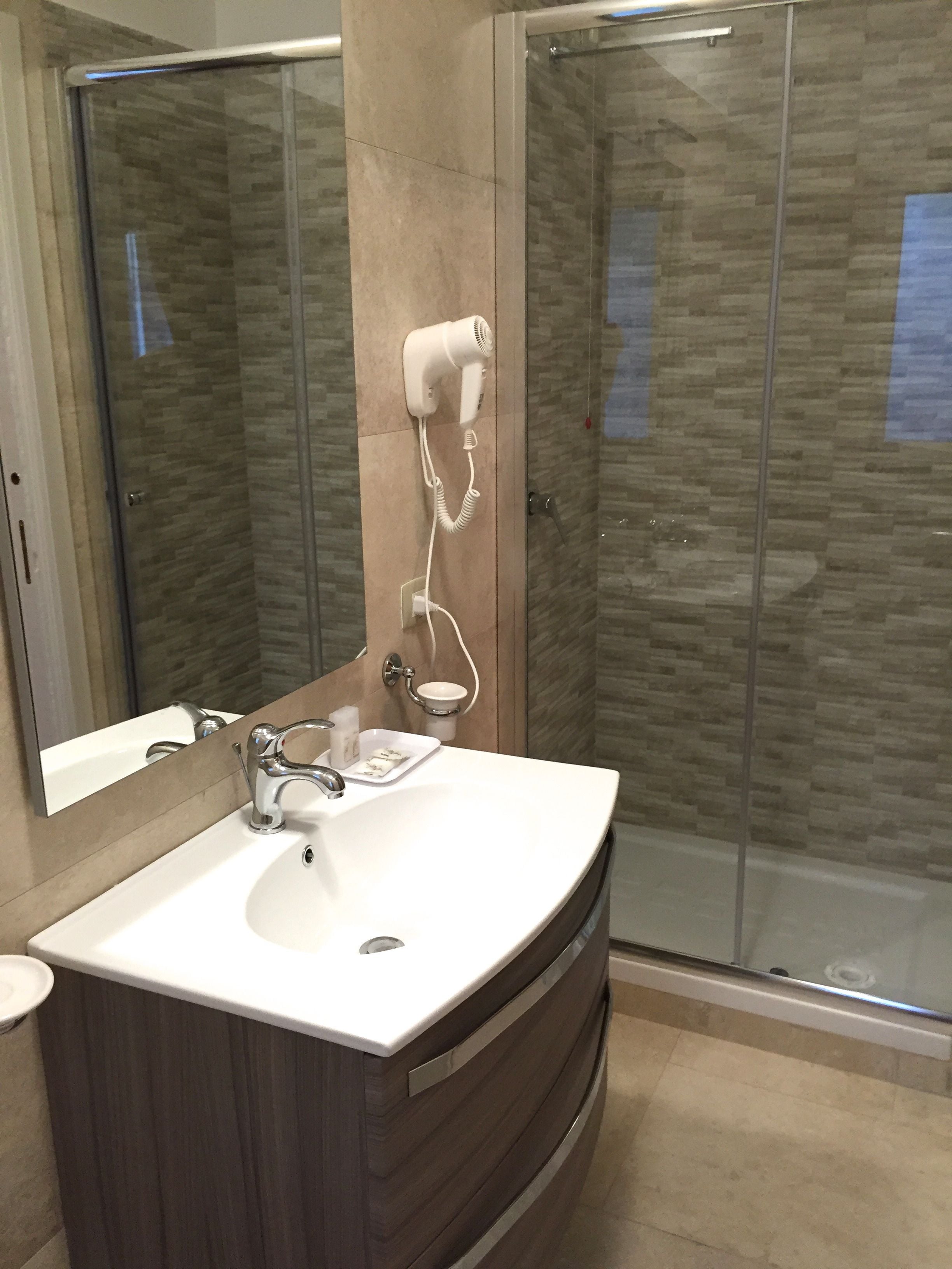 A bathroom with a white sink, a mirror, a hairdryer mounted on the wall, and a glass shower enclosure.