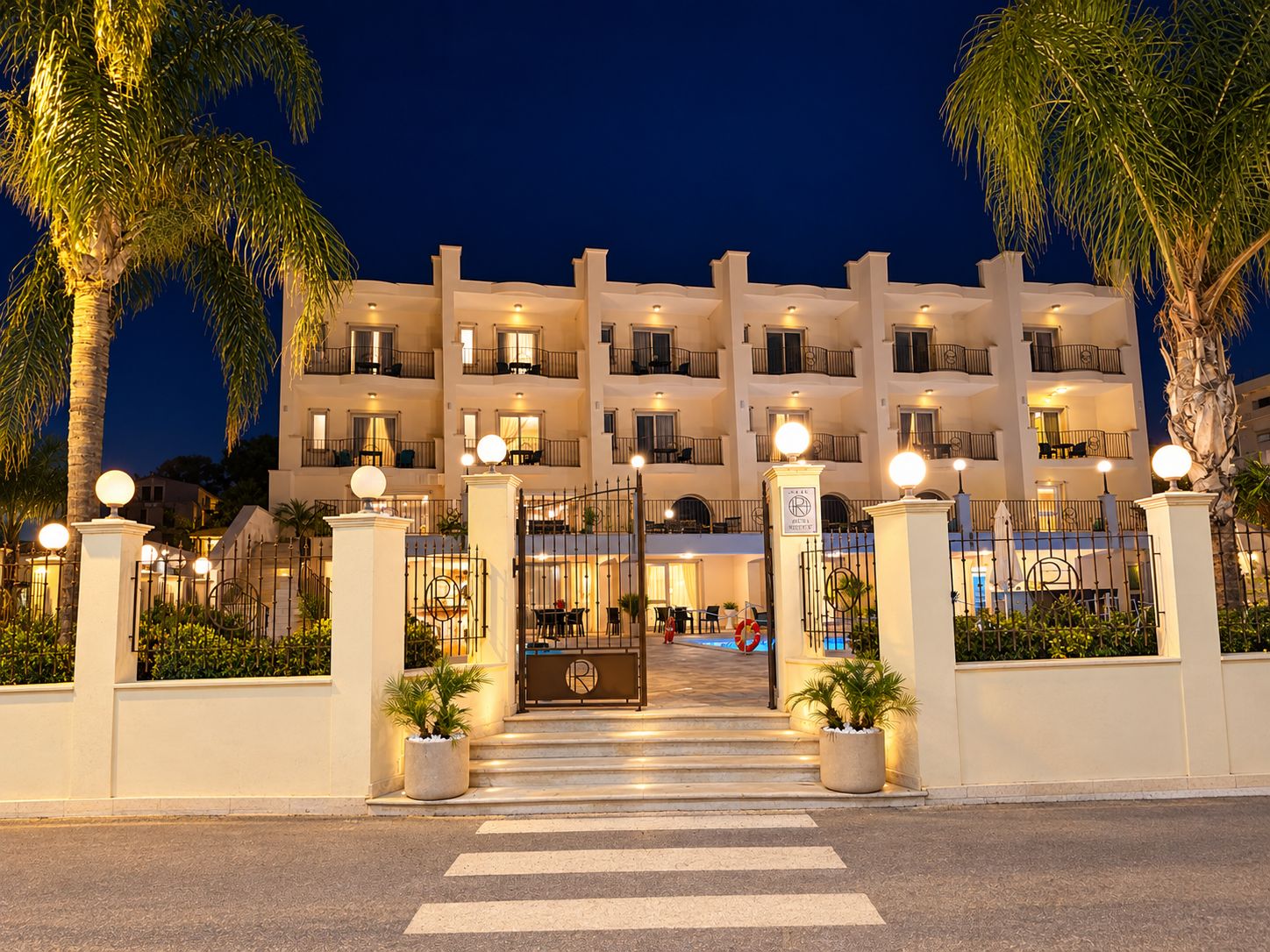 Un hotel su più piani con ingresso chiuso da cancello, palme e una facciata ben illuminata.