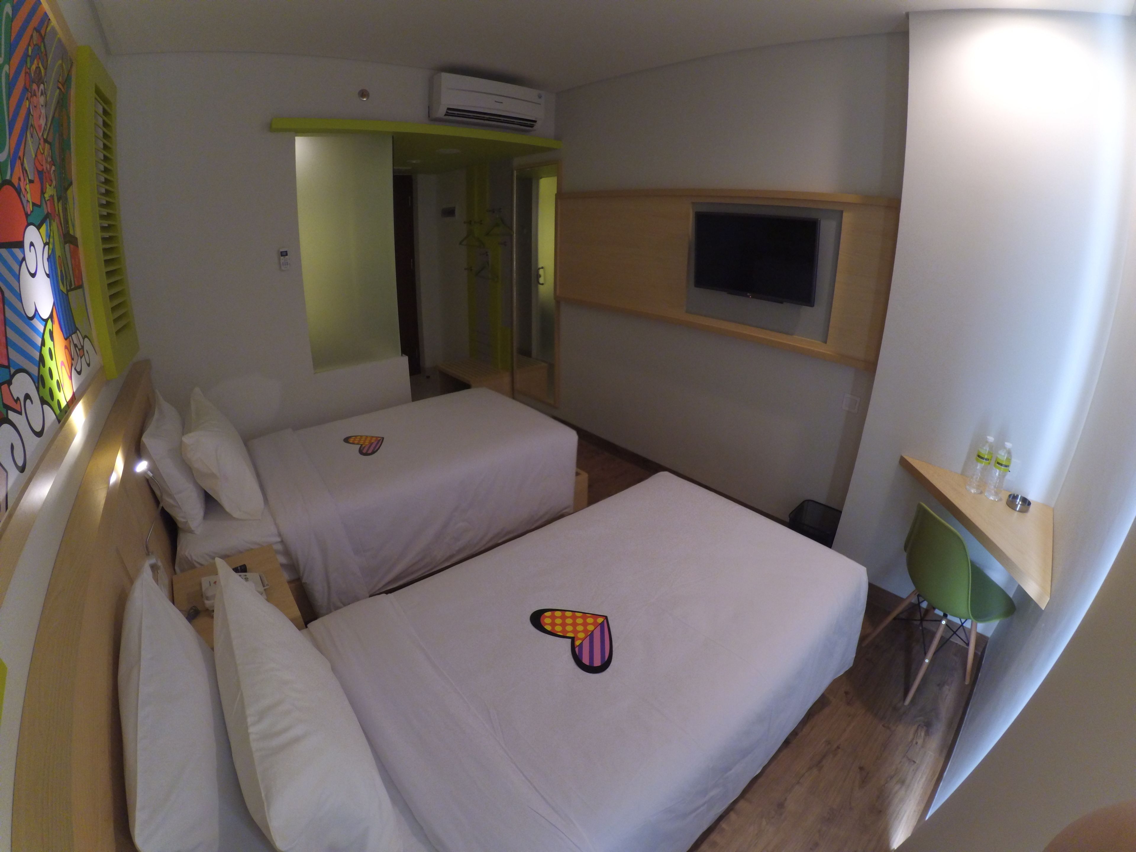 A hotel room with two beds, a TV mounted on the wall, and a small shelf with water bottles.