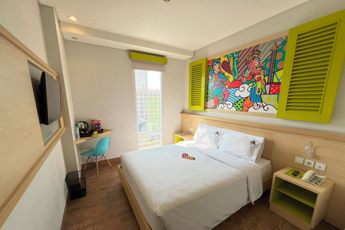 A hotel room with a bed, a desk, a chair, a TV, a window, and a colorful painting on the wall.