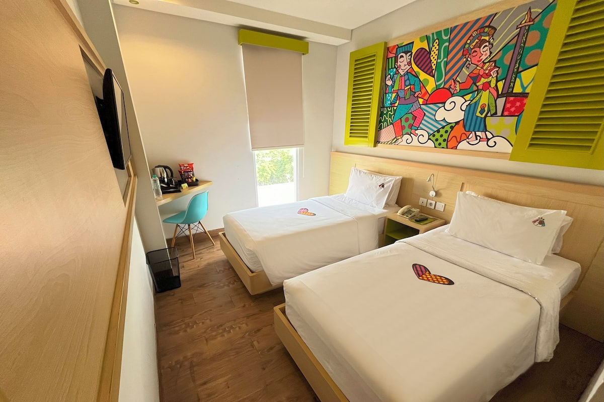 A hotel room with two beds, a desk with a chair, a window, and a colorful painting on the wall.