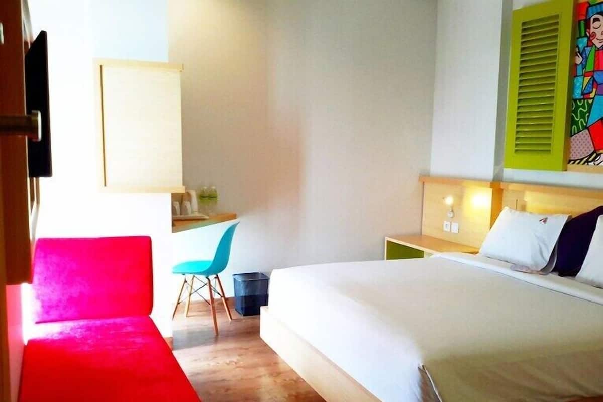 A hotel room with a bed, a desk with a chair, a small table, and a colorful artwork on the wall.