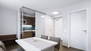 Una habitación moderna y minimalista con una mesa y sillas blancas, y una cocina visible a través de un separador de vidrio. Se observa una puerta cerrada.