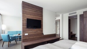 Habitación de hotel moderna con una cama grande, un panel de madera decorativo, un televisor de pantalla plana montado encima y un escritorio con una silla, además de un armario.