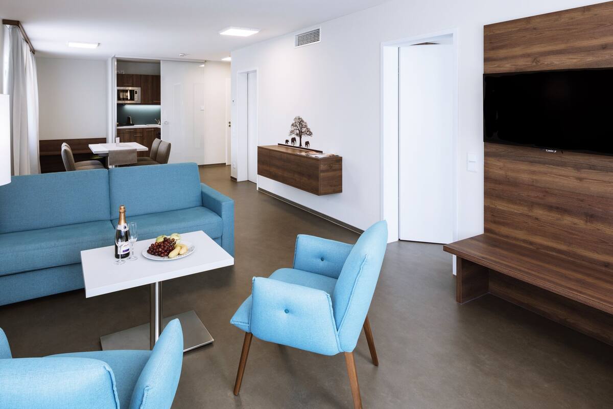 A modern living room with a blue sofa, a small table with a bottle and glasses, and a TV mounted on a wooden wall.