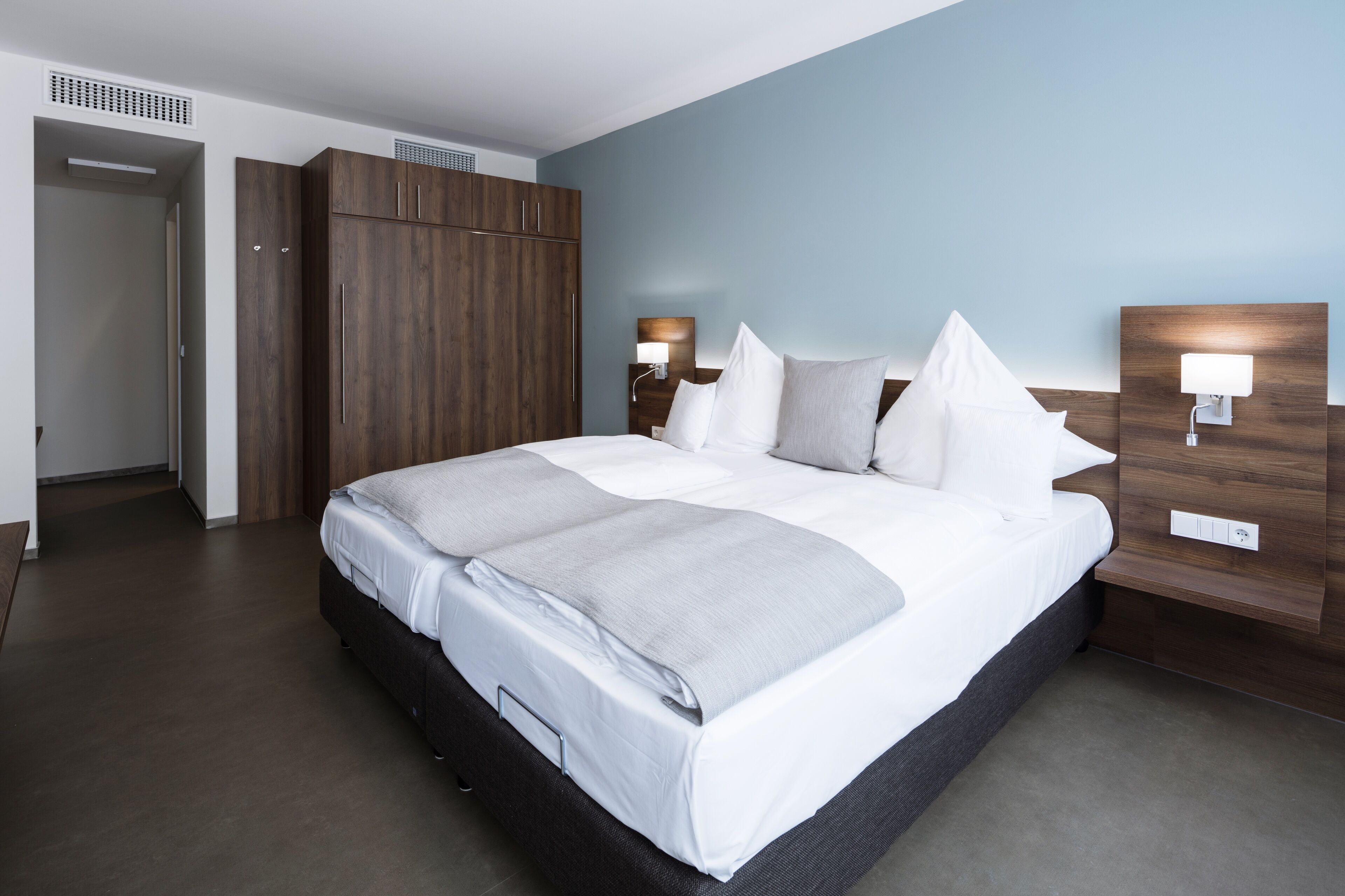 A modern hotel room with a large bed, wooden wardrobe, bedside lamps, and a neutral color scheme.
