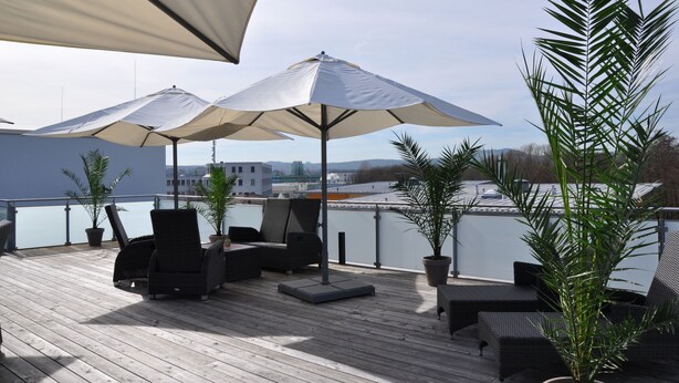 Terraza en la azotea con dos sombrillas grandes, mobiliario de mimbre, plantas en macetas y vistas panorámicas de la ciudad.