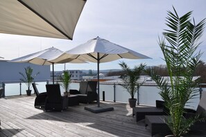 A rooftop terrace with two large umbrellas, wicker furniture, potted plants, and a view of the cityscape. - Apart Hotel FirstBoarding Bayreuth (Bayreuth)