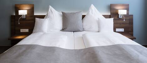Una cama bien hecha con sábanas blancas y una manta gris, con dos mesitas de noche a cada lado y lámparas fijadas a la pared.