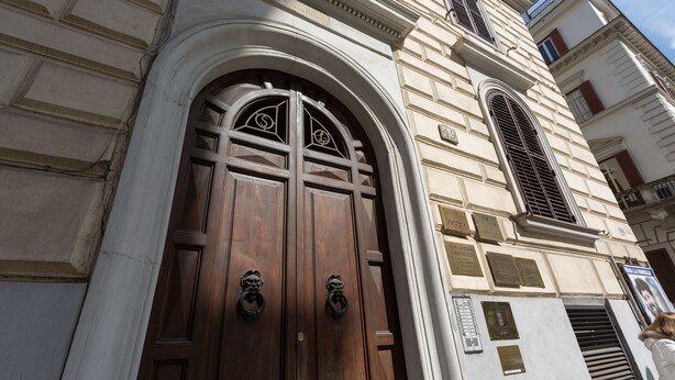 A large, ornate wooden door with a decorative arch and two lion head handles, set in a classical building facade with stonework and a window with shutters.