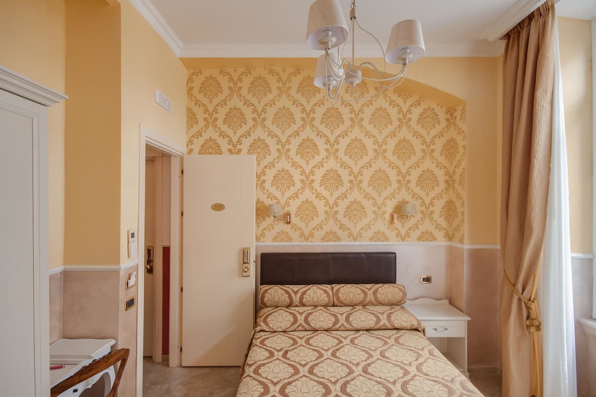 A bedroom with a patterned bedspread, a bedside table, a nightstand, a wardrobe, and a chandelier.