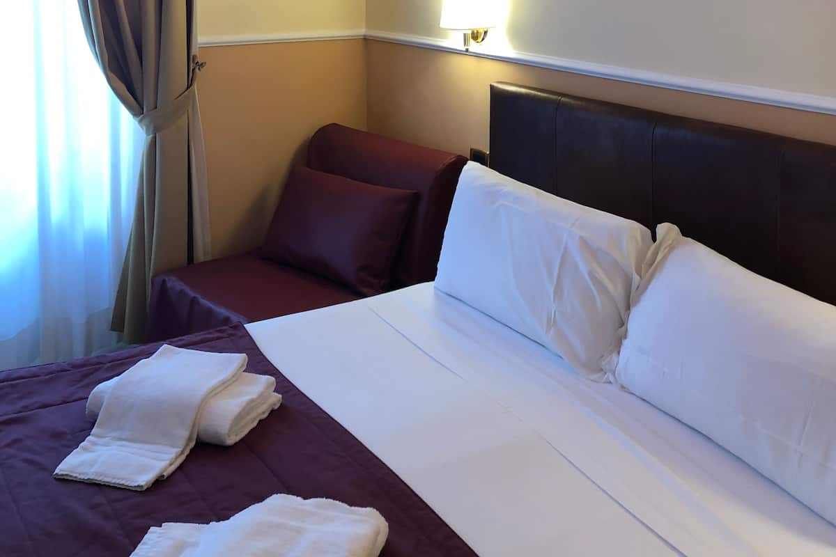 A hotel room with a large bed, a headboard, a painting of a cityscape, and a window with curtains.