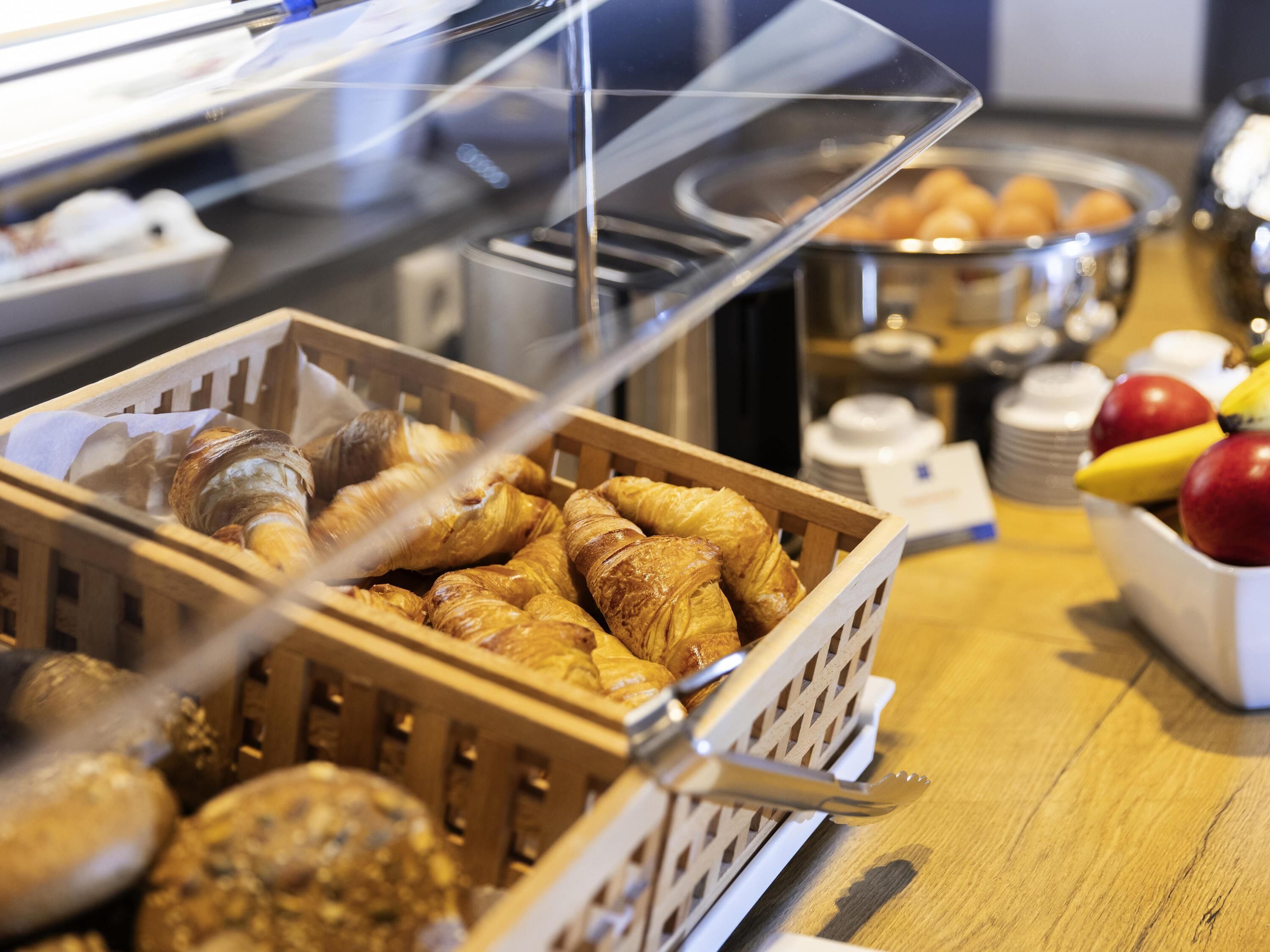 Un buffet avec un panier en bois rempli de viennoiseries, un saladier de fruits contenant des pommes et des bananes, et un couvercle en plastique transparent recouvrant le présentoir de nourriture.