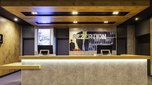 A reception desk with a wooden finish, a large wall-mounted screen, and a modern ceiling design. - ibis budget Ludwigsburg (Ludwigsburg)